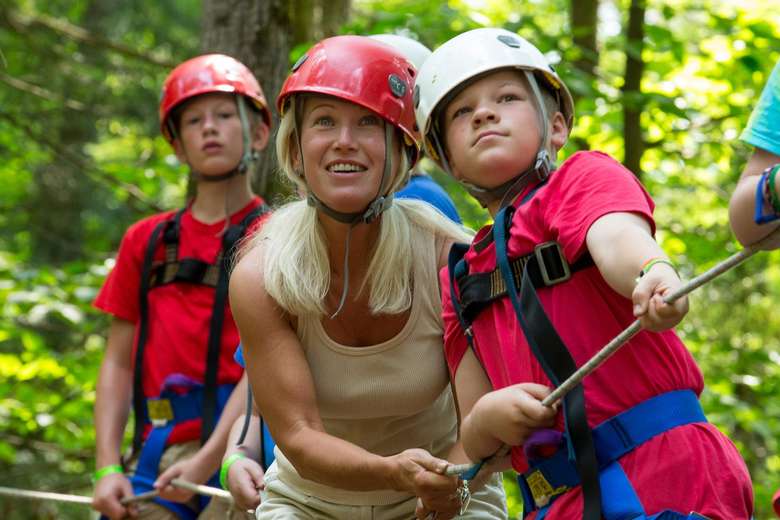 Female counselor in a red helmet helping kids in red and white helmets through a ropes course